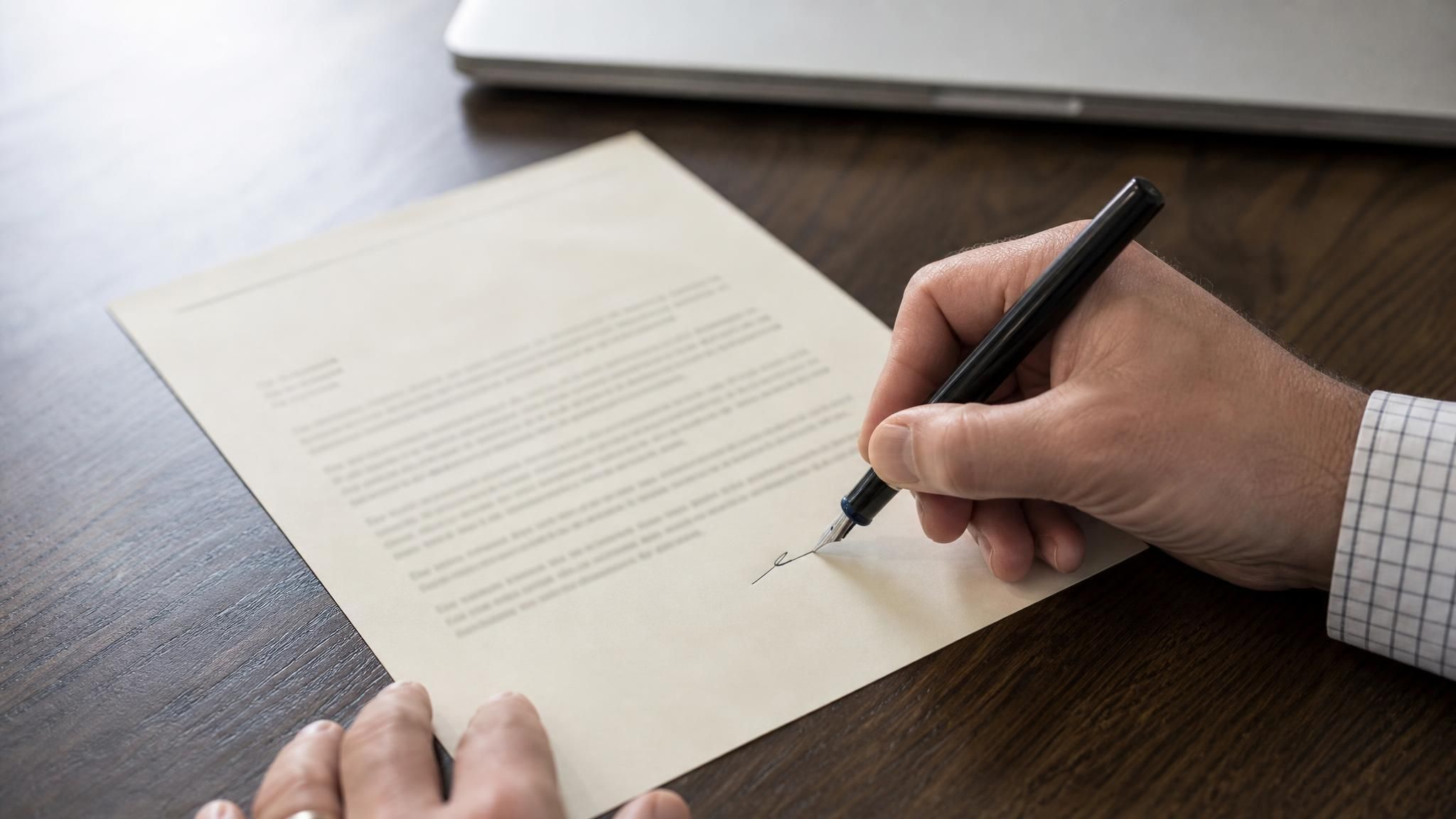 A hand signs a printed AI letter of recommendation on institutional letterhead beside a closed laptop, the final step before sending.