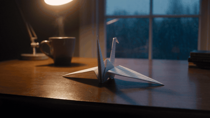 A white origami crane on a walnut desk lifts its wings and takes off past a steaming mug and a desk lamp, with a rainy blue-hour window behind, generated by Seedance 2.5 through Claude Code
