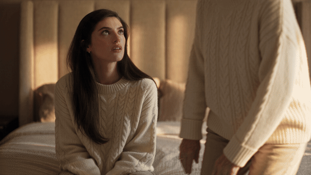 Hug your younger self AI flipped: a 25-year-old woman and her 80-year-old self in matching cream sweaters sit on a bed in warm window light, hug, and laugh with their foreheads almost touching