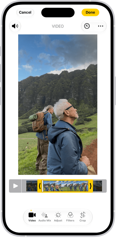 Screenshot of the Photos app Edit screen on iPhone showing a video of two hikers below green cliffs, with yellow trim handles on the frame viewer under the video and the Video, Audio Mix, Adjust, Filters and Crop buttons along the bottom Screenshot from Apple's iPhone User Guide, Trim video length, adjust speed, and edit audio on iPhone.