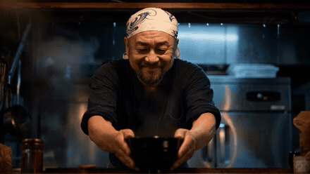 Smiling chef wearing a bandana serves a steaming black bowl