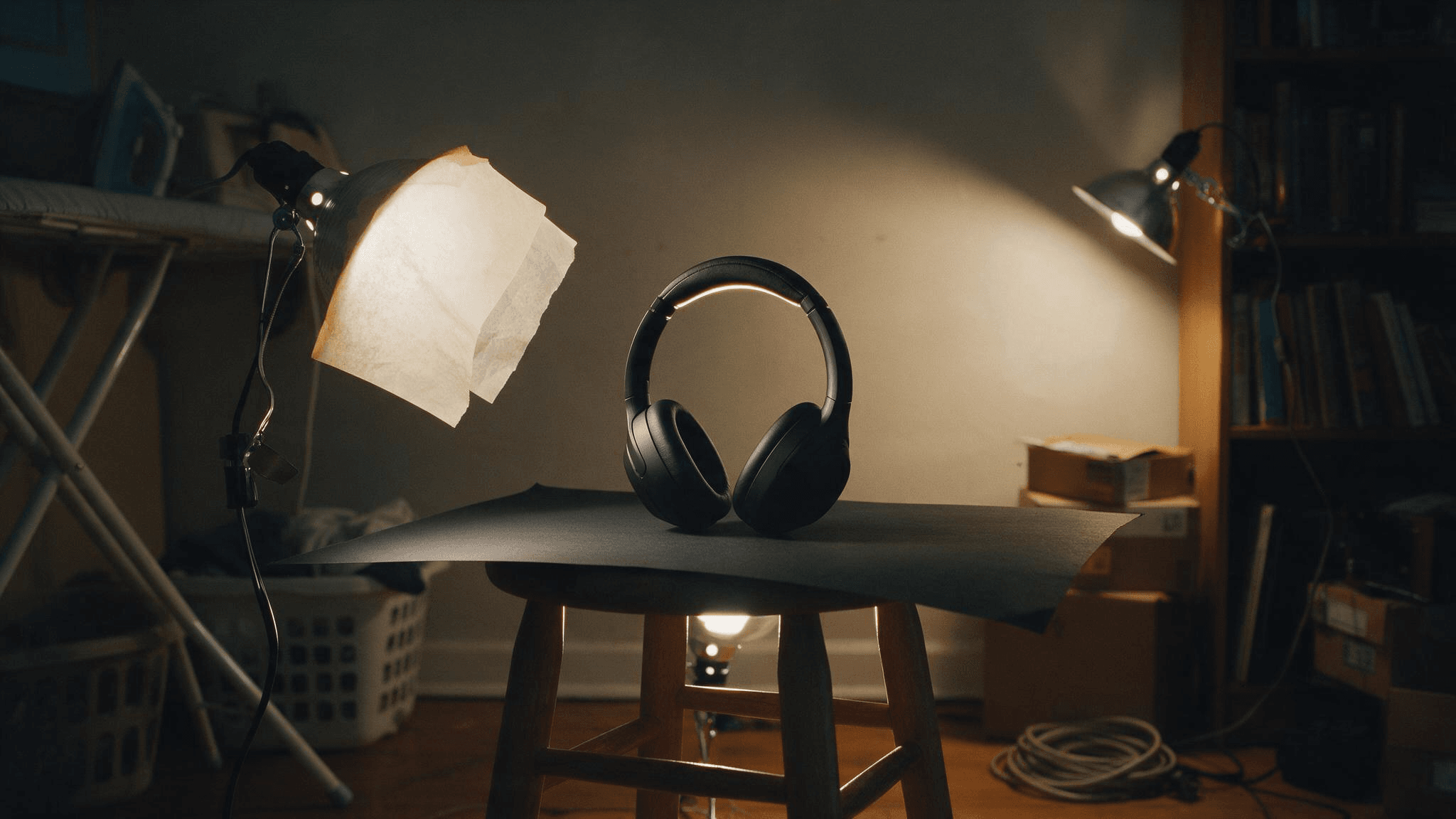 Makeshift home product studio at night: matte black over-ear headphones on a wooden stool covered with black poster board, lit by a clamp lamp with baking parchment over it on the left and a lamp clipped to a bookshelf on the right, in a dim spare room with an ironing board, a laundry basket, cardboard boxes and a coiled extension cord