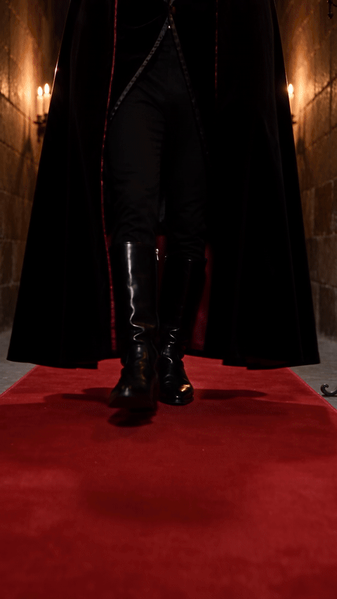 Vampire ramalama costume walk made with Seedance 2.5 from a black-and-white portrait, showing boots on a red carpet, a tilt up to a long-haired man in a velvet jacket and lace jabot, and a crimson-lined cape sweep