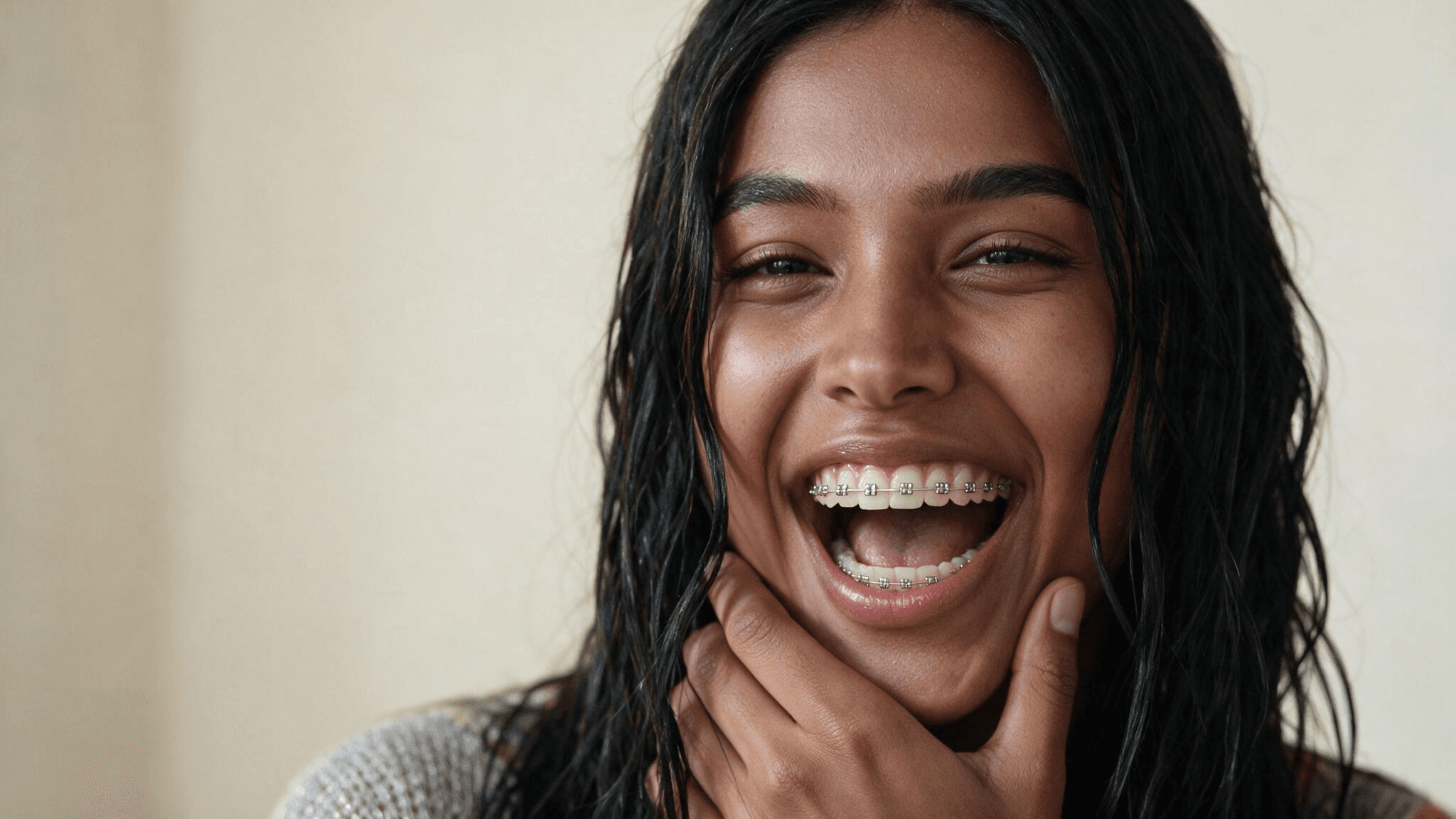 Braces filter CapCut trend, a close-up of a woman with long dark wavy hair laughing with metal braces on her teeth and one hand resting on her jaw