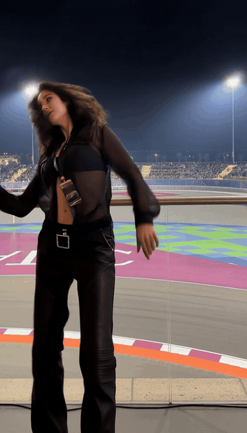 Animated clip of an AI-generated woman in a sheer black blouse and black trousers dancing in front of a floodlit night racetrack, pointing one arm up, wrapping both arms over her head and covering her mouth