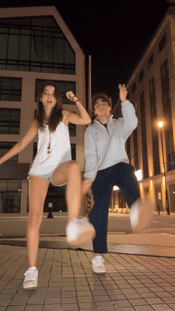 Animated clip of an AI-generated couple, a woman in a white camisole and denim shorts and a man in a grey half-zip sweatshirt, dancing in sync on a city street at night with wide low stances and palms-up shrugs